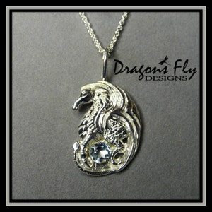 Op de afbeelding: Zilveren hanger met een gedetailleerd drakenontwerp met een blauwe edelsteen in het midden. De hanger is aan een zilveren ketting en is gelabeld "Dragons Fly Designs".