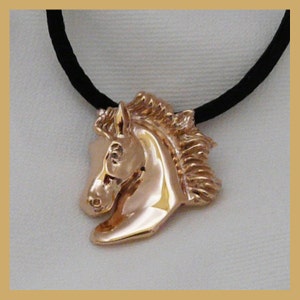 Pode incluir: Um colar com um pingente de cabe&ccedil;a de cavalo dourado em um cord&atilde;o preto. A cabe&ccedil;a do cavalo est&aacute; voltada para a esquerda e tem uma crina detalhada.