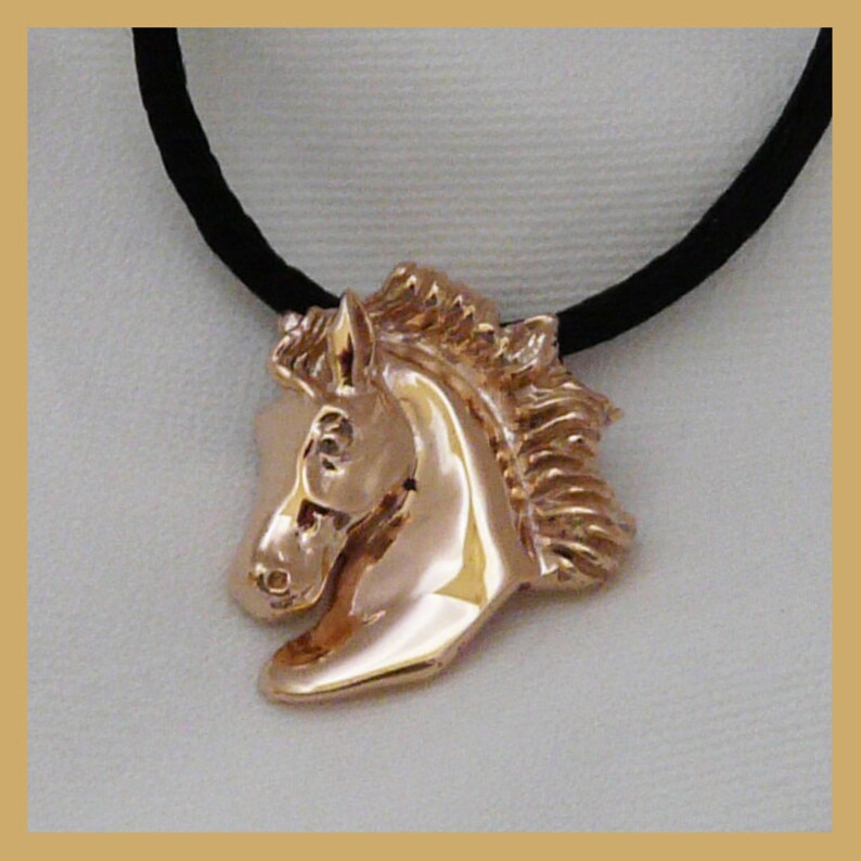 Pode incluir: Um colar com um pingente de cabe&ccedil;a de cavalo dourado em um cord&atilde;o preto. A cabe&ccedil;a do cavalo est&aacute; voltada para a esquerda e tem uma crina detalhada.