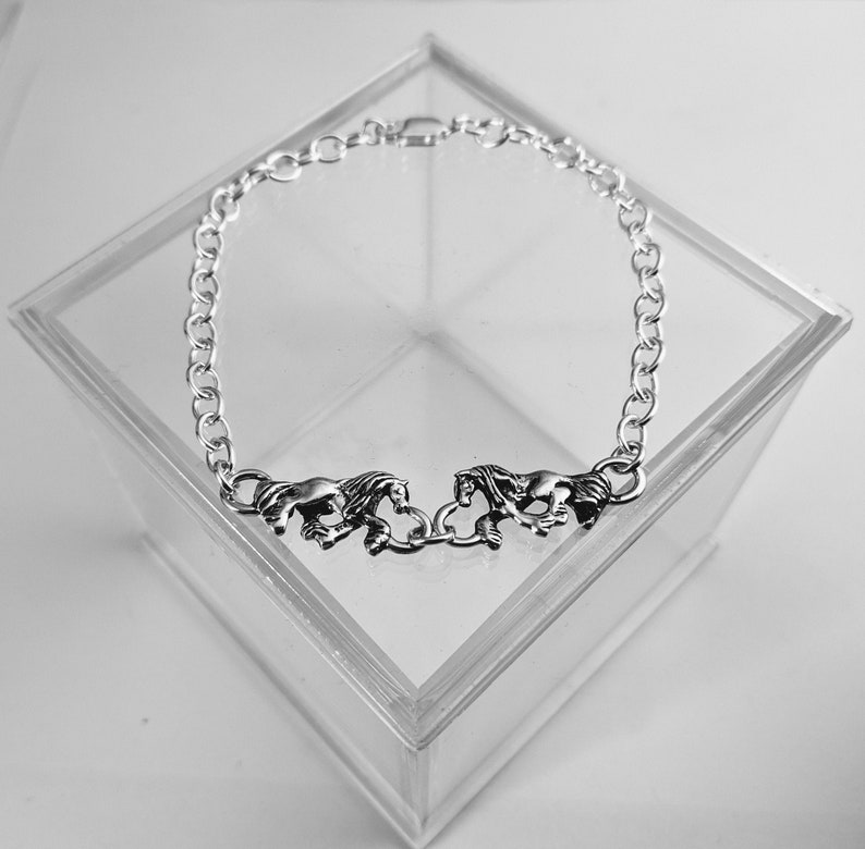 May include: Silver chain bracelet with three horse charms. The bracelet is displayed in a clear plastic box.