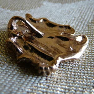Pode incluir: Um pingente de metal dourado em forma de folha estilizada. O pingente tem um la&ccedil;o na parte superior para prender a uma corrente ou cord&atilde;o.