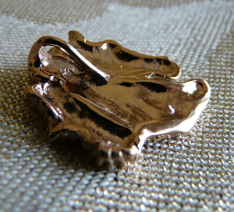Pode incluir: Um pingente de metal dourado em forma de folha estilizada. O pingente tem um la&ccedil;o na parte superior para prender a uma corrente ou cord&atilde;o.