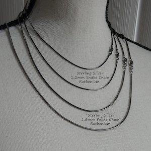K&ouml;nnte beinhalten: F&uuml;nf silberne Schlangenketten in verschiedenen St&auml;rken auf einer wei&szlig;en Puppe. Die Ketten sind mit "Sterling Silver 1.2mm Snake Chain Ruthenium" und "Sterling Silver 1.6mm Snake Chain Ruthenium" beschriftet.