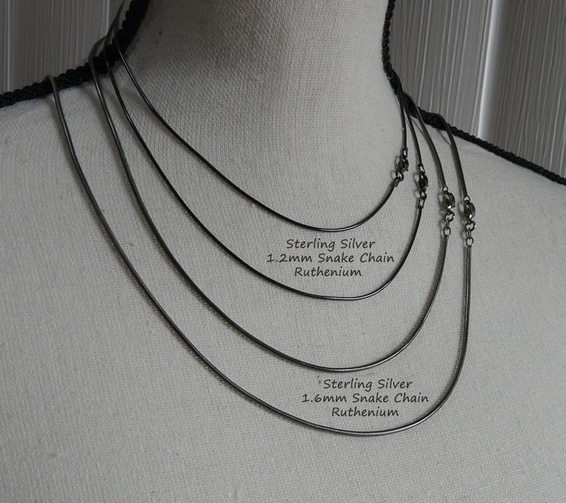 K&ouml;nnte beinhalten: F&uuml;nf silberne Schlangenketten in verschiedenen St&auml;rken auf einer wei&szlig;en Puppe. Die Ketten sind mit "Sterling Silver 1.2mm Snake Chain Ruthenium" und "Sterling Silver 1.6mm Snake Chain Ruthenium" beschriftet.