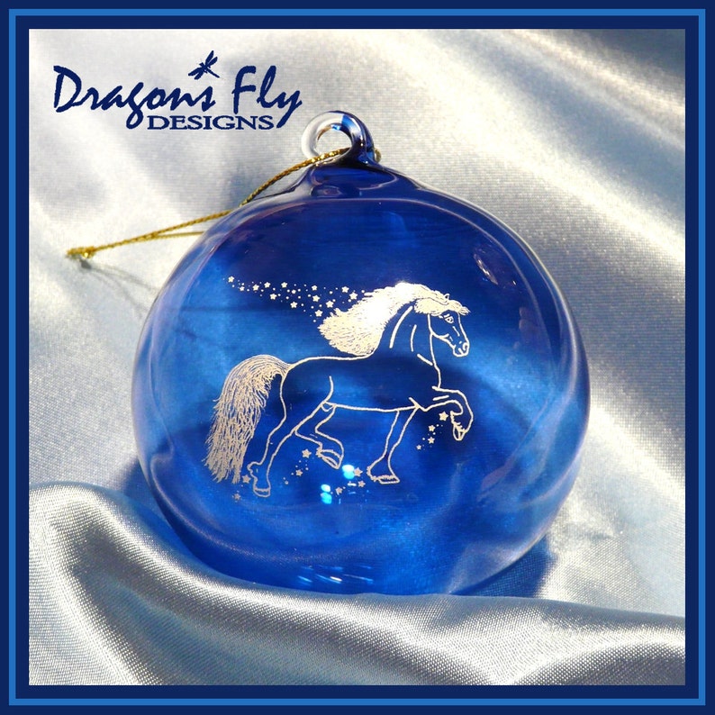 May include: A blue glass Christmas ornament with a white horse design and gold glitter. The ornament is decorated with a white horse with a flowing mane and tail, and the text "Dragons Fly Designs" is visible in the top left corner.