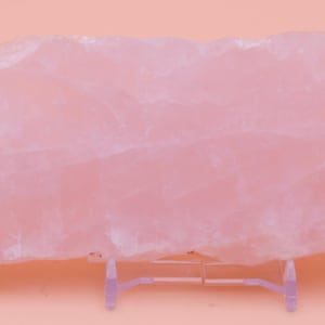 May include: A large, raw rose quartz crystal slab, in a soft pink colour. The stone has a rough, natural texture and is mounted on a clear plastic stand. The crystal's surface shows subtle variations in colour and veining.