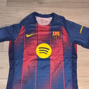 May include: A blue and red FC Barcelona soccer jersey featuring a yellow Nike swoosh, the FC Barcelona crest, and a yellow Spotify logo on the front. The fabric displays multiple black ink signatures across the front panels and the word AMBILIC.