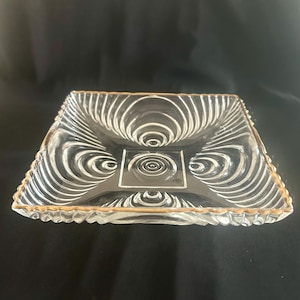May include: Clear glass square dish with a gold rim and a decorative swirl pattern. The dish has a square center with a spiral design and a scalloped edge. The glass is transparent, allowing the background to show through.