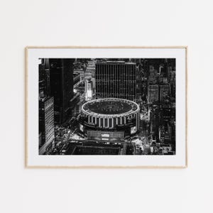 May include: A black and white aerial photograph of Madison Square Garden in New York City, framed in a light wood border. The image captures the circular roof of the arena surrounded by dense city skyscrapers and illuminated urban streets at night.