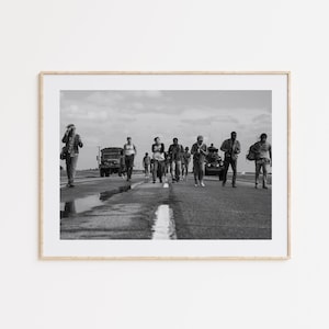May include: A black and white framed photograph depicting a group of people walking along a paved road. In the background, a military-style truck and a tank are visible. The individuals are dressed in casual clothing and carry various bags and gear.