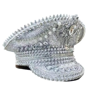 Op de afbeelding: Zilveren hoed in militaire stijl met een met studs bezette rand en kroon. De hoed is versierd met pailletten, imitatieparels en grote, heldere strass-steentjes. De hoed heeft een bruine rand en is ontworpen voor een glamoureuze look.