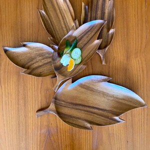 May include: Set of four wooden leaf-shaped serving trays. The trays are made of dark brown wood with a natural grain pattern. The trays are arranged in a flower shape with the center tray containing small pieces of fruit and cucumber.
