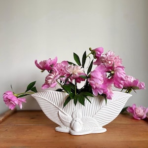 May include: A white ceramic vase shaped like a wide, elongated leaf with a central embossed motif, filled with a bouquet of vibrant pink peony flowers with green stems and leaves, displaying a textured, ribbed surface across the exterior.