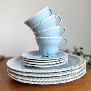 May include: A stack of four light blue teacups with white trim, balanced atop a stack of matching saucers. The teacups have a classic design with curved handles. The plates and cups are arranged on a wooden surface.