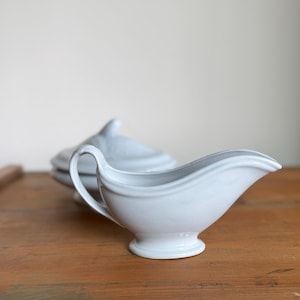 May include: A light blue ceramic gravy boat with a handle and a small, round base. A matching covered serving dish is in the background. The items are on a wooden surface.