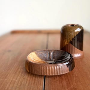 May include: A ceramic ashtray and vase set. The ashtray is round with a ribbed exterior and a brown, tan, and black color scheme. The vase is cylindrical with a similar color palette and a diagonal color block design.