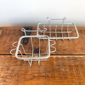 May include: Two vintage metal soap dishes. The soap dishes are made of silver-colored wire and have a rectangular shape with a grid pattern. One dish has a square shape with decorative side elements. The dishes are displayed on a wooden surface.