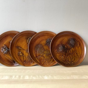 May include: Four round wooden plates with raised floral designs. The plates feature different floral patterns, including sunflowers, irises, and other flowers. The plates are a warm brown color and have a wood grain texture.