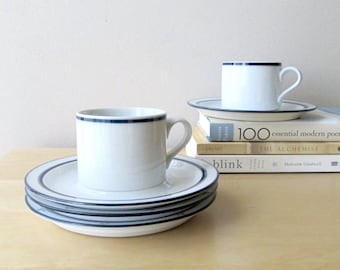 May include: Two white ceramic coffee cups with blue trim, one on a stack of four matching saucers, the other on a single saucer. The cups and saucers are on a light wood surface. A stack of books is in the background.