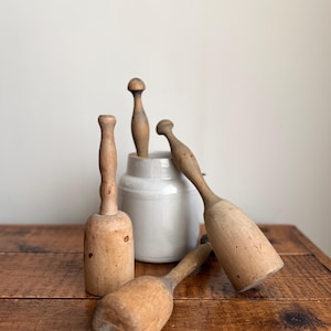 May include: A collection of vintage wooden muddlers and a small ceramic crock on a rustic wooden surface. The muddlers vary in shape and size, with some featuring a rounded head and others a cylindrical design. The crock is a light gray color.