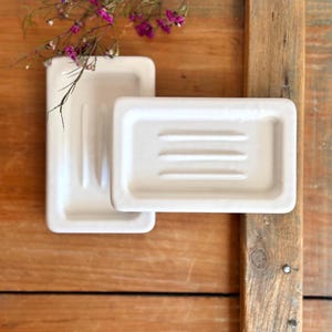 May include: Two off-white rectangular soap dishes, one with three parallel ridges, are displayed on a wooden surface. A sprig of purple flowers adds a touch of color. The soap dishes are a simple, elegant design.