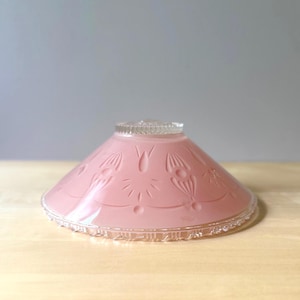 May include: A conical glass lamp shade in a soft pink hue featuring a molded floral pattern around the exterior. The shade has a clear, textured glass rim at the base and a matching clear, ridged glass collar at the top opening for mounting.