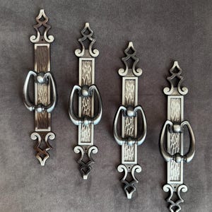 May include: Four ornate metal door pulls with a dark silver finish. Each pull features a rectangular textured center, a curved handle, and decorative top and bottom elements. The pulls are arranged vertically against a brown background.