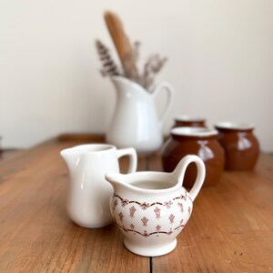 May include: A collection of ceramic cream pitchers and small pots on a wooden surface. Two white pitchers are in the foreground, one plain and one with a decorative red pattern. In the background, a white pitcher holds dried flowers and a baguette.
