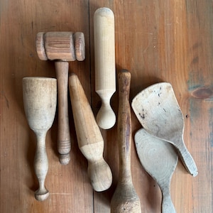 May include: A collection of antique wooden kitchen utensils, including a mallet, rolling pin, and various spoons and paddles. The items are arranged on a wooden surface, showcasing their natural wood grain and aged appearance.