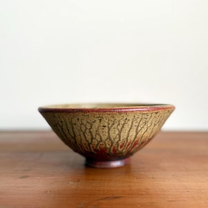 May include: A ceramic bowl featuring a wide, flared rim and a tapered base. The exterior surface displays a textured, crackled glaze pattern in shades of beige and tan, transitioning into a deep, solid red glaze toward the bottom of the bowl.