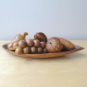 May include: A wooden fruit bowl filled with carved wooden fruit. The bowl is oval-shaped and contains a pear, grapes, a star fruit, and other fruits. The fruit and bowl are a light brown color.