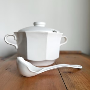 May include: A white ceramic soup tureen with a matching lid and a soup ladle. The tureen has a geometric design and two handles. The ladle is resting on a wooden surface.