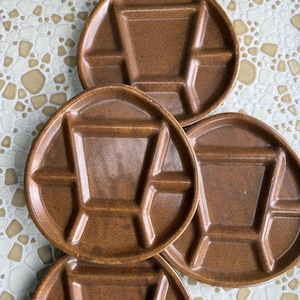 May include: A set of four brown ceramic divided plates. Each plate has a circular shape with a central square and four surrounding compartments. The plates are arranged on a patterned surface.