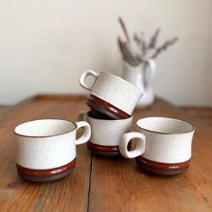 May include: Four ceramic mugs with a speckled white finish and a brown band around the base. The mugs have a simple, rounded handle and are stacked on a wooden surface. The mugs are a neutral color and would fit in any kitchen.