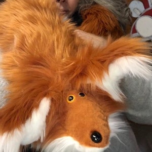 May include: A plush fox toy with orange fur, white accents, and black eyes. The toy is being held by a person.