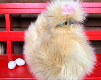 May include: A fluffy, cream-coloured plush chicken with long, soft faux fur, a small grey beak, black bead eyes, and a light blue accent near the eye. A pale pink fabric flower is attached to the top of its head. Two small white eggs sit nearby.