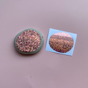 May include: A close-up of a round, iridescent sticker with a copper glitter design. The sticker is on a light blue background. The sticker is also shown adhered to a light green, round, textured surface.