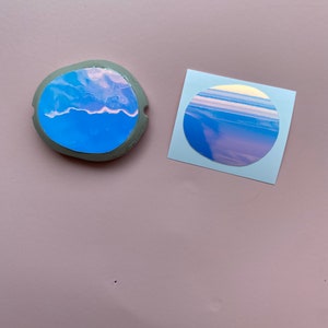 May include: Two round stickers with a holographic sky design. One sticker is on a gray round surface and the other is on a blue square surface.
