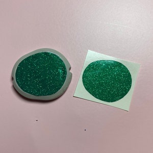 May include: Two green glitter circles, one on a white background and one on a gray background. The circles are the same size and shape.