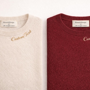 Pode incluir: Dois suéteres de gola redonda, um creme e um bordô, são exibidos lado a lado. Cada suéter tem o texto "Custom Text" bordado em fio dourado. O suéter creme tem uma etiqueta que diz "RosesAllDesign".