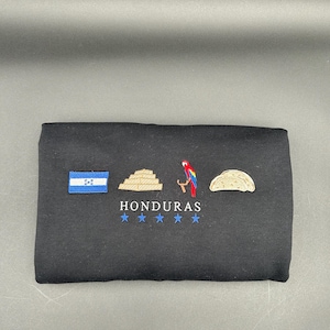 May include: Black t-shirt with embroidered images of the Honduran flag, a Mayan pyramid, a red macaw, and a Honduran pupusa. The text "HONDURAS" is embroidered below the images, with five stars underneath.
