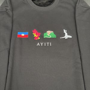 May include: Dark gray sweatshirt with embroidered designs. Features a flag, flowers, a mountain, and a figure. The word "AYITI" is embroidered below the designs. A casual, comfortable garment.