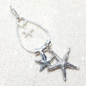 Pode incluir: Pingente de prata com uma moldura em forma de lágrima com três pingentes de estrela do mar pendurados. As estrelas do mar são texturizadas e têm um design ligeiramente elevado.