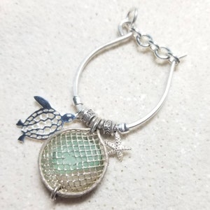 May include: A silver-toned bracelet featuring a sea-themed design. The bracelet includes a turtle charm, a round cage with sea glass, and a starfish charm. The bracelet has a toggle clasp and is displayed on a white surface.
