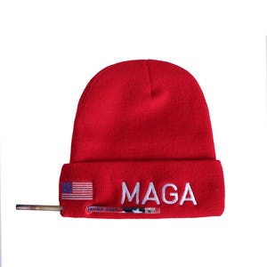 May include: Red knit beanie with a white embroidered "MAGA" and a small American flag on the side. The beanie also has a gold-colored pin with the text "TRUMP 2024" on it.