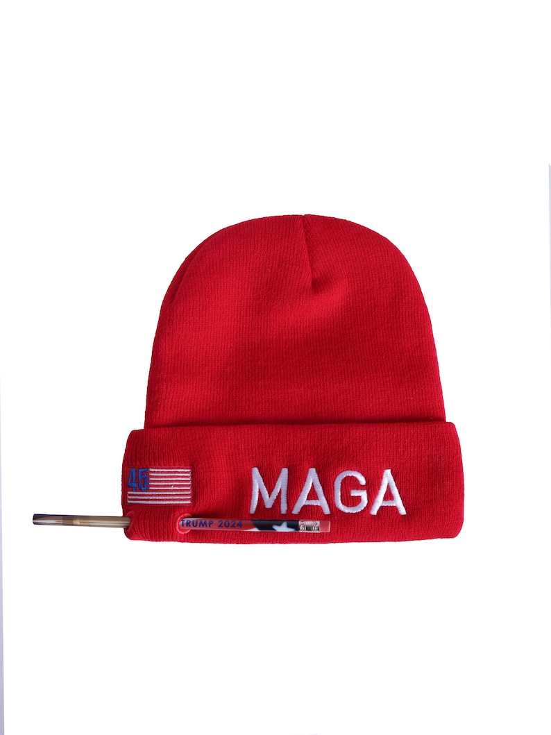 May include: Red knit beanie with a white embroidered "MAGA" and a small American flag on the side. The beanie also has a gold-colored pin with the text "TRUMP 2024" on it.