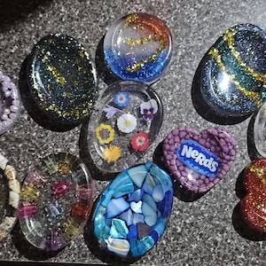 May include: A collection of resin art pieces in various shapes, including ovals, hearts, and circles. Some pieces feature glitter, dried flowers, and seashells. One piece has the word "Nerds" on it.