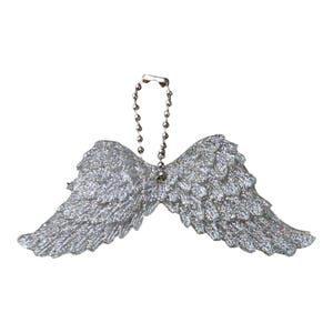 May include: A silver glitter-covered angel wing ornament featuring detailed feather textures and a metallic ball chain loop attached at the center top for hanging. The ornament has a symmetrical design with pointed tips extending outward on both sides.