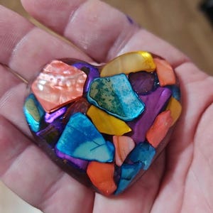 May include: A heart-shaped decorative object with a mosaic design. The mosaic is composed of iridescent, colorful pieces in shades of orange, blue, yellow, and purple. The heart is held in a hand.