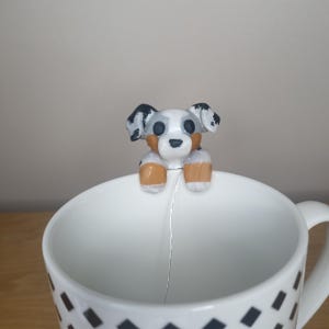 Peut inclure: Une tasse en céramique blanche avec un motif en diamant noir et blanc. Une petite figurine de chien peinte à la main est perchée sur le bord de la tasse.